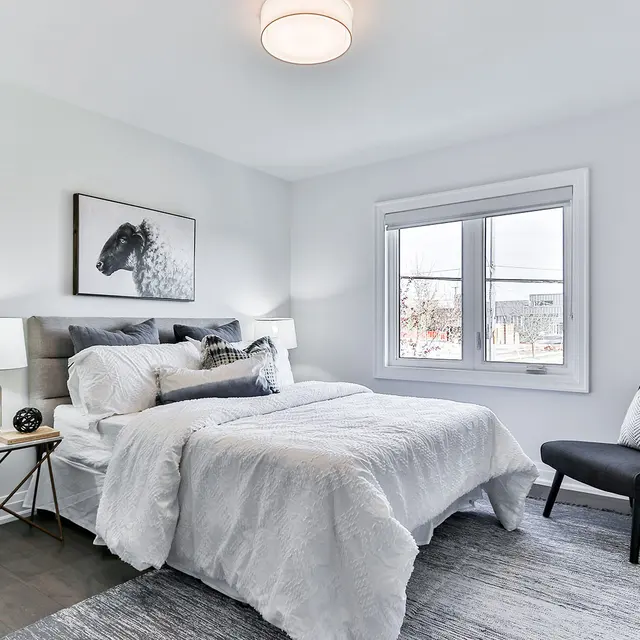 A modern bedroom featuring a double bed, decorative pillows, a lamp, and a small chair beside a window with natural light.