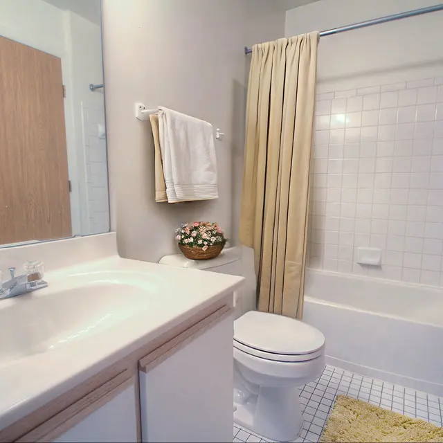 A clean bathroom featuring a white sink with a vanity, a bathtub with a beige curtain, and a small flower arrangement on the counter. Brightly lit with neutral walls and tiled flooring.