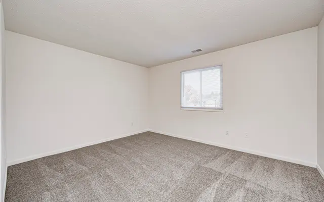 A spacious empty room with beige carpet and white walls. There is a single window allowing natural light to enter.