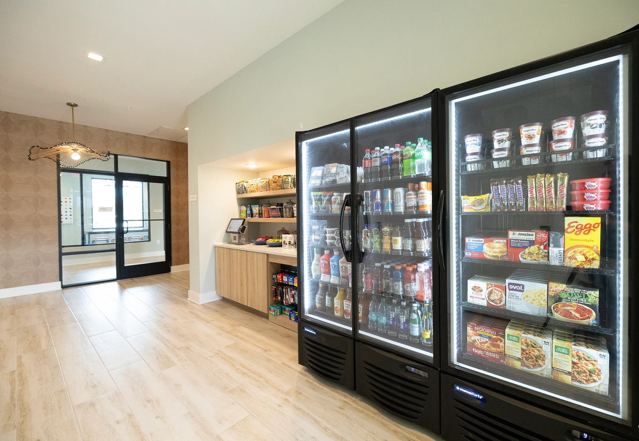 Modern Snack Area A brightly lit snack area featuring glass-fronted refrigerators filled with drinks and packaged food items. On one side, there's a wooden shelf with assorted snacks and a digital kiosk. The room has light-colored flooring and a doorway visible in the background.