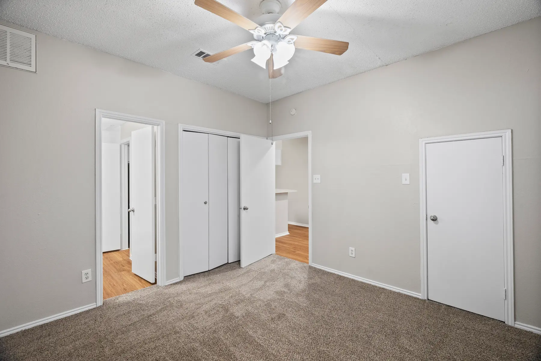 A spacious bedroom with light gray walls and carpeted flooring. It features a ceiling fan with wooden blades, two sets of white closet doors, and two doors leading to different areas. The room is well-lit and appears organized and tidy.