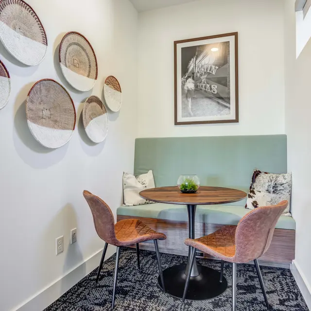 A cozy corner meeting space featuring a round wooden table surrounded by two brown chairs. The walls are adorned with circular wall art and there is a framed picture above the table. A small plant centerpiece is on the table, creating a warm and inviting atmosphere.