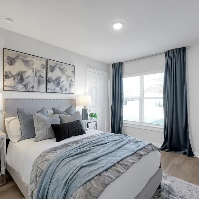A well-decorated modern bedroom featuring a bed with gray and white bedding, two bedside lamps on metallic side tables, and large windows with blue drapes.