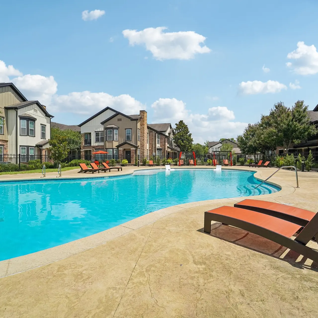 Community Pool Area A swimming pool surrounded by lounge chairs and trees, with apartment buildings in the background on a sunny day.