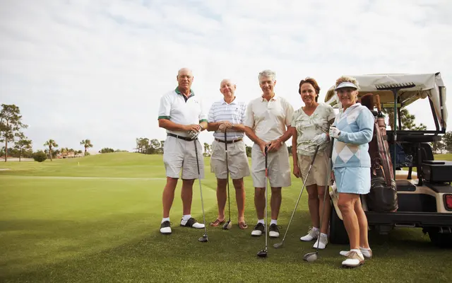 Seniors Golfing Together A group of five older adults standing on a golf course, smiling at the camera. They are dressed in golf attire, holding golf clubs, with a golf cart parked nearby.