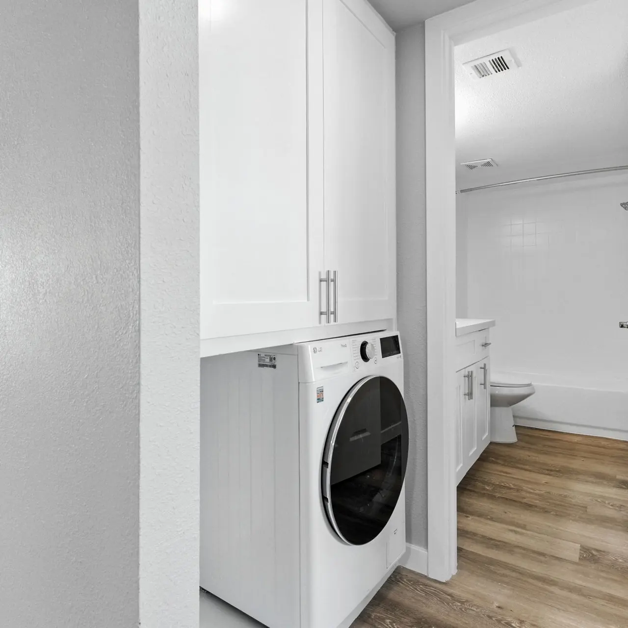 Modern Laundry Room with Bathroom Access A modern laundry room with a front-loading washing machine and white cabinets, leading toward a bathroom with a tub. The floor is wooden and the walls are painted light gray.