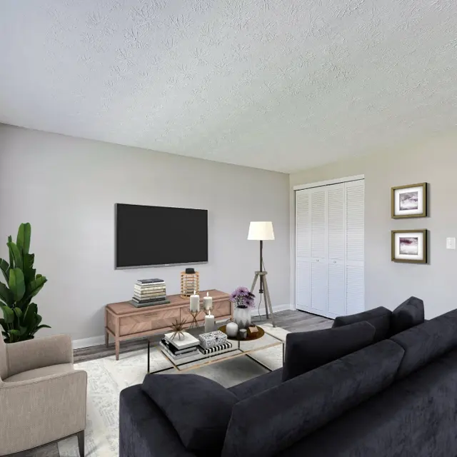 A modern living room featuring a black sofa, beige armchair, wooden coffee table, and flat-screen TV mounted on the wall. The room has large windows allowing natural light, a potted plant, and light-colored walls.