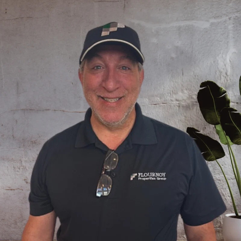 A smiling man wearing a dark shirt and glasses, standing in front of a light grey background with green plants.