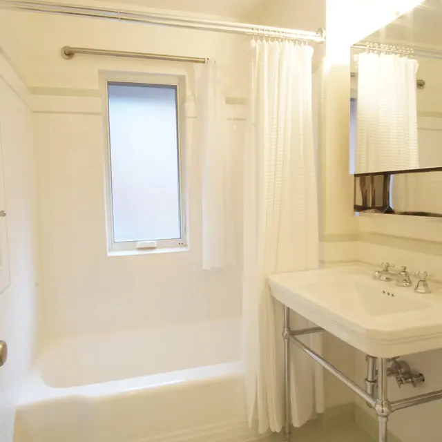A clean, minimalist bathroom featuring a shower/tub combination, a sink with a modern faucet, and a large mirror. The bathroom is well-lit with natural light coming from a window.