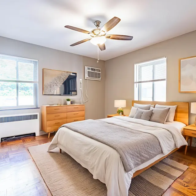 A modern bedroom with a wooden bed, nightstands, and a dresser. The room features large windows, a mounted TV, and a ceiling fan. Decor includes plants and artwork on the walls.