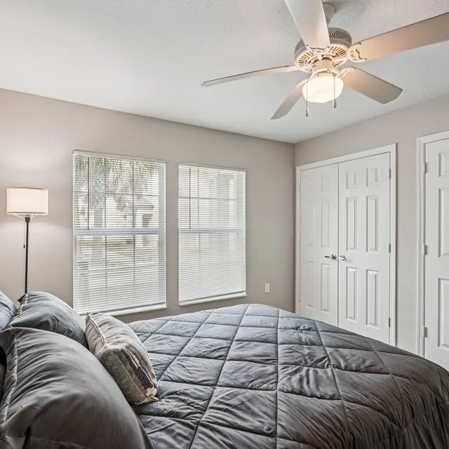 A cozy bedroom featuring a large bed with dark bedding, a ceiling fan, and two windows allowing natural light. The room includes a standing lamp beside the bed and double closet doors on one wall.