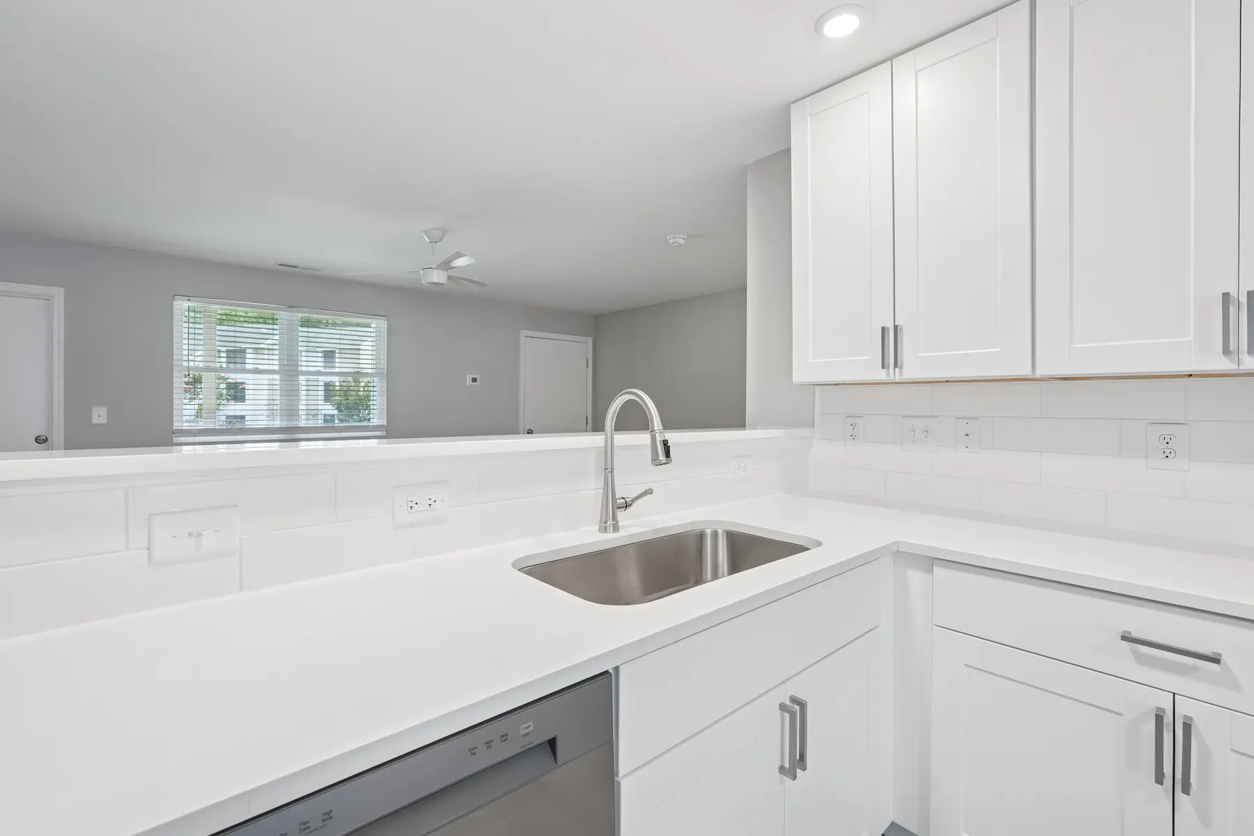 A modern kitchen featuring white cabinets, a stainless steel sink, and a clean countertop with a view of an adjoining room through a window.