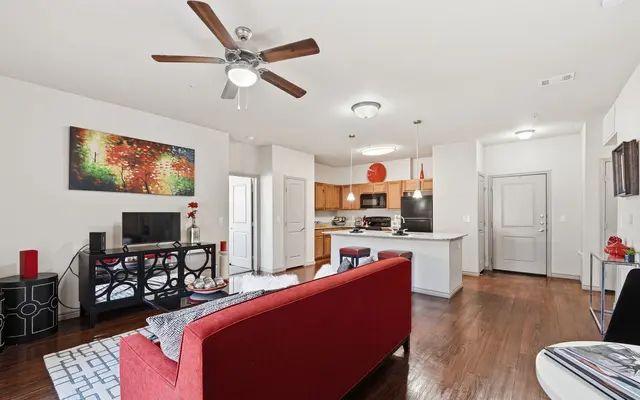 A stylish modern living room featuring a red sofa, a coffee table with decor, and an open kitchen area with wooden cabinets. There's a ceiling fan and colorful artwork on the walls.