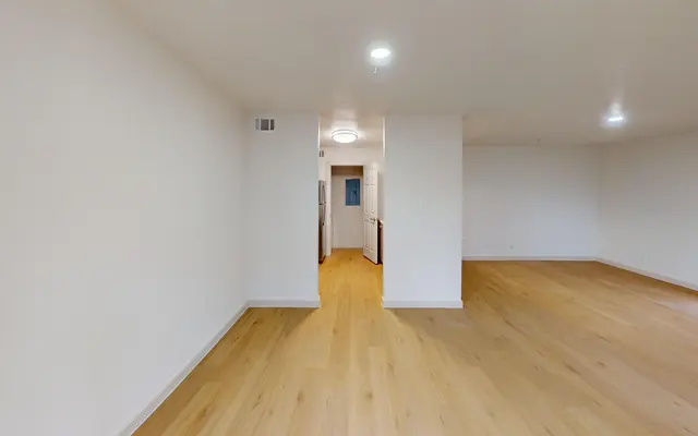 The Gleason A spacious and bright living room with light-colored walls and wooden flooring, featuring a pathway leading to a kitchen area in the background.