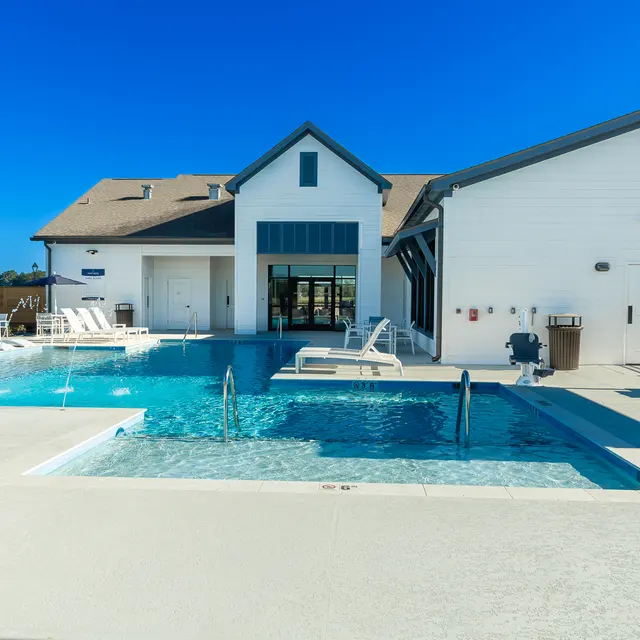 A modern pool area featuring a large swimming pool with lounge chairs, a shaded umbrella, and a contemporary building in the background under a clear blue sky.