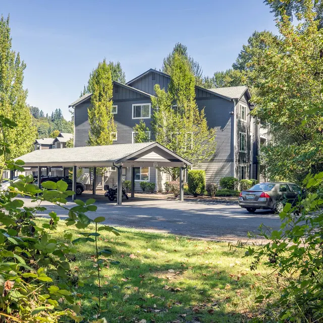 A two-story building surrounded by trees and a parking area, seen from a grassy area with bushes in the foreground.