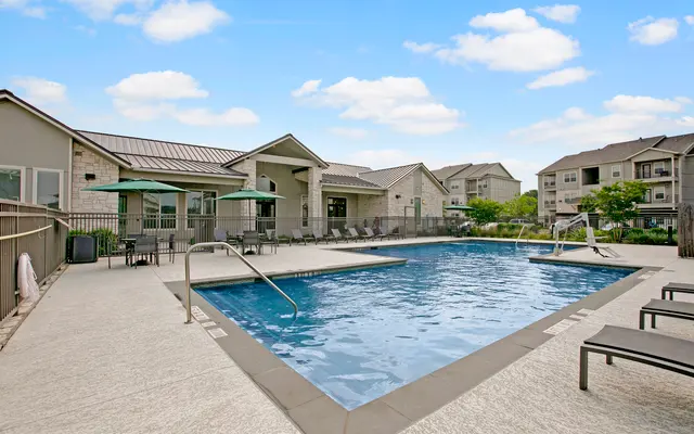 A serene apartment complex pool area with clear blue water, surrounded by lounge chairs and shaded seating under umbrellas.