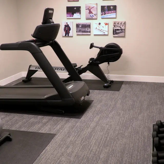 A spacious home gym with various exercise equipment including a treadmill, stationary bike, and an elliptical machine, alongside dumbbells and a stability ball. The walls feature framed photographs related to fitness.