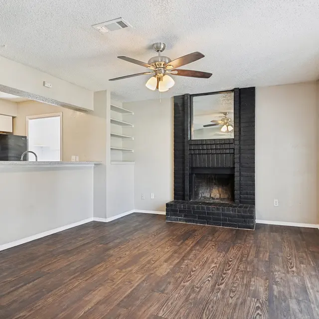 A spacious living room featuring dark hardwood flooring, a ceiling fan with lights, and a black brick fireplace. There is a partial wall separating the living area from a kitchen space, which is partially visible in the background.