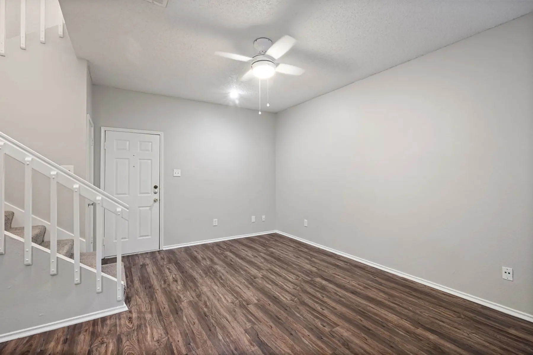 An empty living room with light wood flooring, a ceiling fan, and a small staircase on the left leading up. The walls are painted a light gray and there is a white door leading outside.