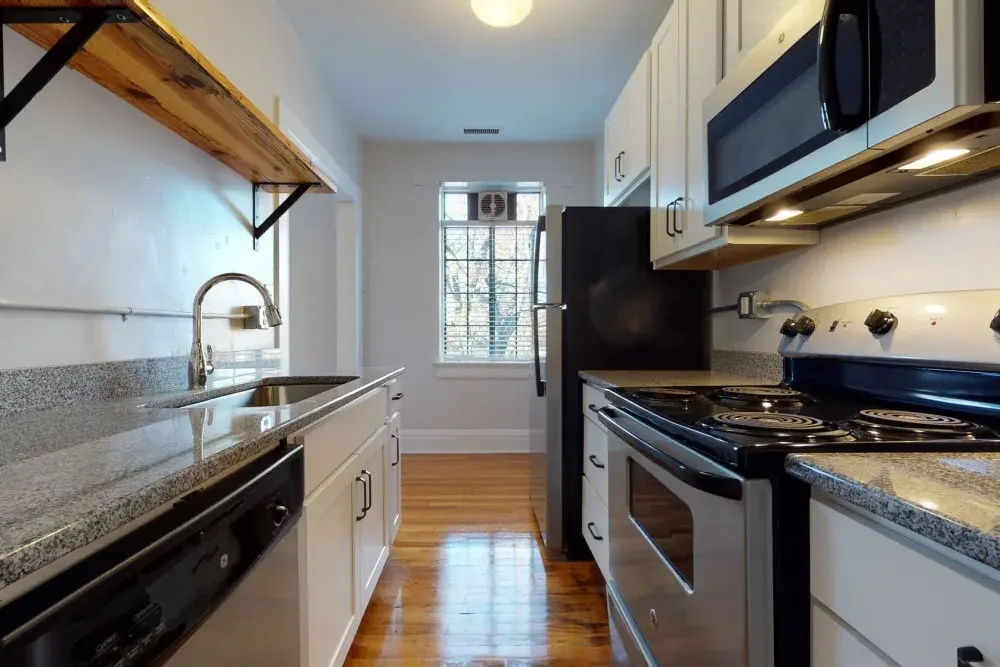 A modern kitchen featuring gray granite countertops, white cabinets, and stainless steel appliances. A wooden shelf lines the wall above the sink, adding a rustic touch.