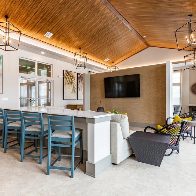 A modern lobby featuring a high ceiling with wood paneling, pendant lights, and a large flat-screen TV. There are bar stools along a kitchen island, a comfortable sofa, and accent chairs. Colorful wall art depicting tropical plants adds a vibrant touch to the decor.