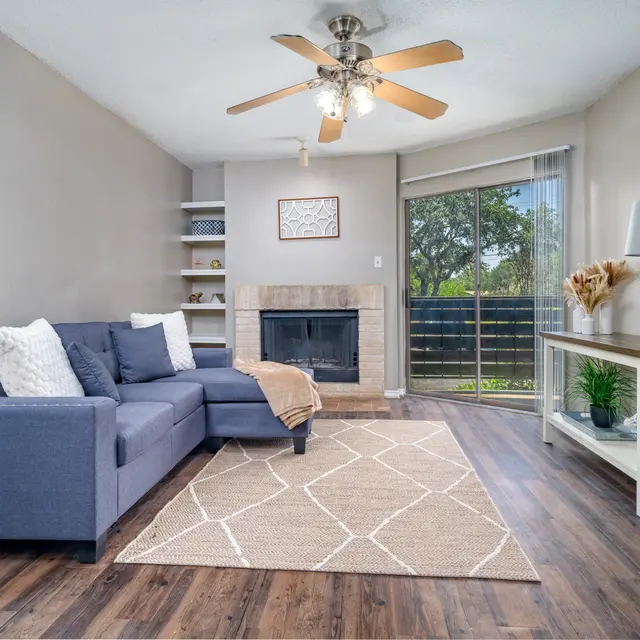 Cozy living room featuring a blue sectional sofa, a beige rug with a geometric pattern, a decorative table with a lamp, and a fireplace. There are large windows that let in natural light and a bookshelf in the background.