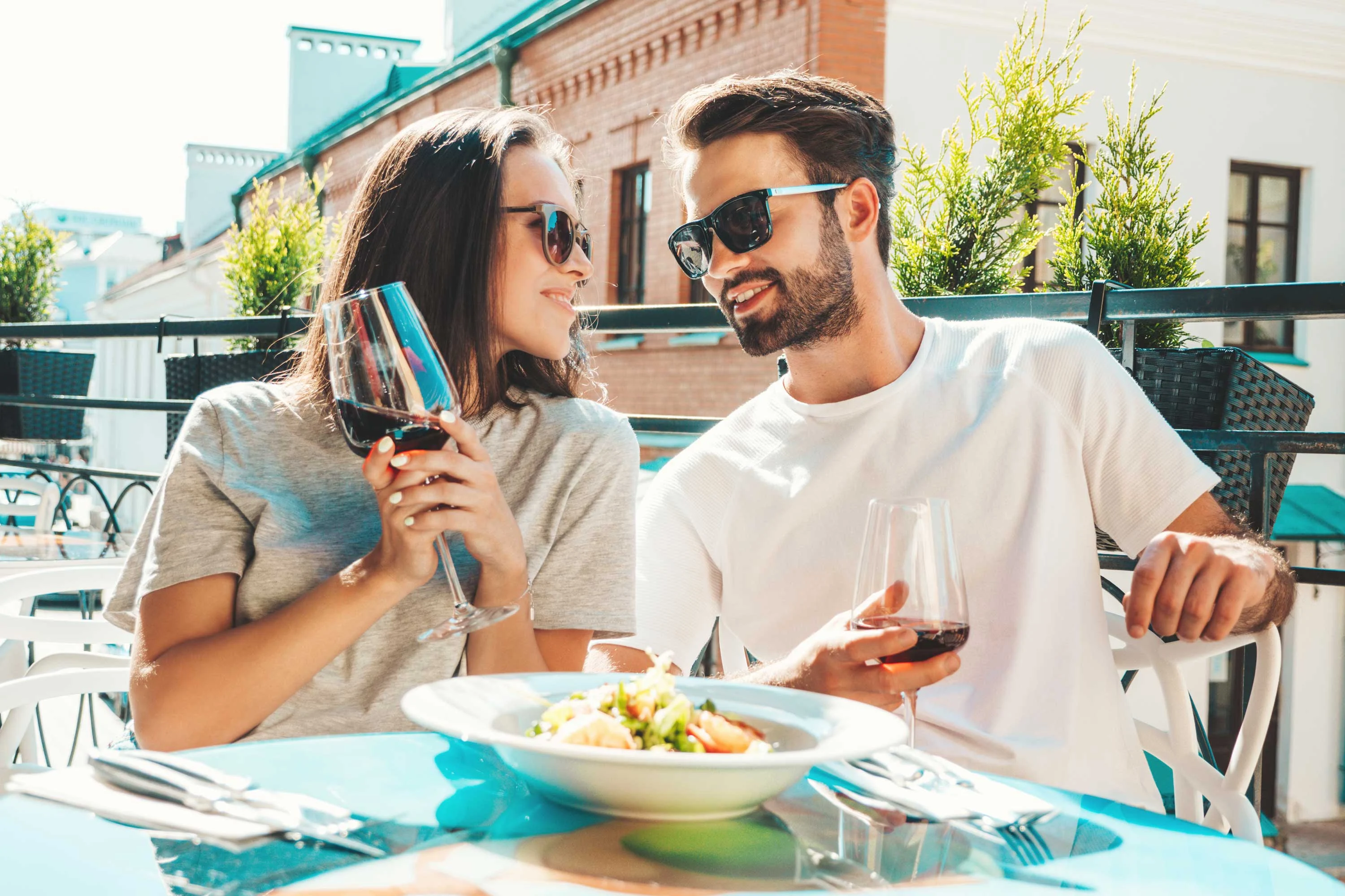 A couple enjoying a meal together on an outdoor terrace, both wearing sunglasses and holding glasses of red wine. They are smiling at each other with a bowl of salad on the table.