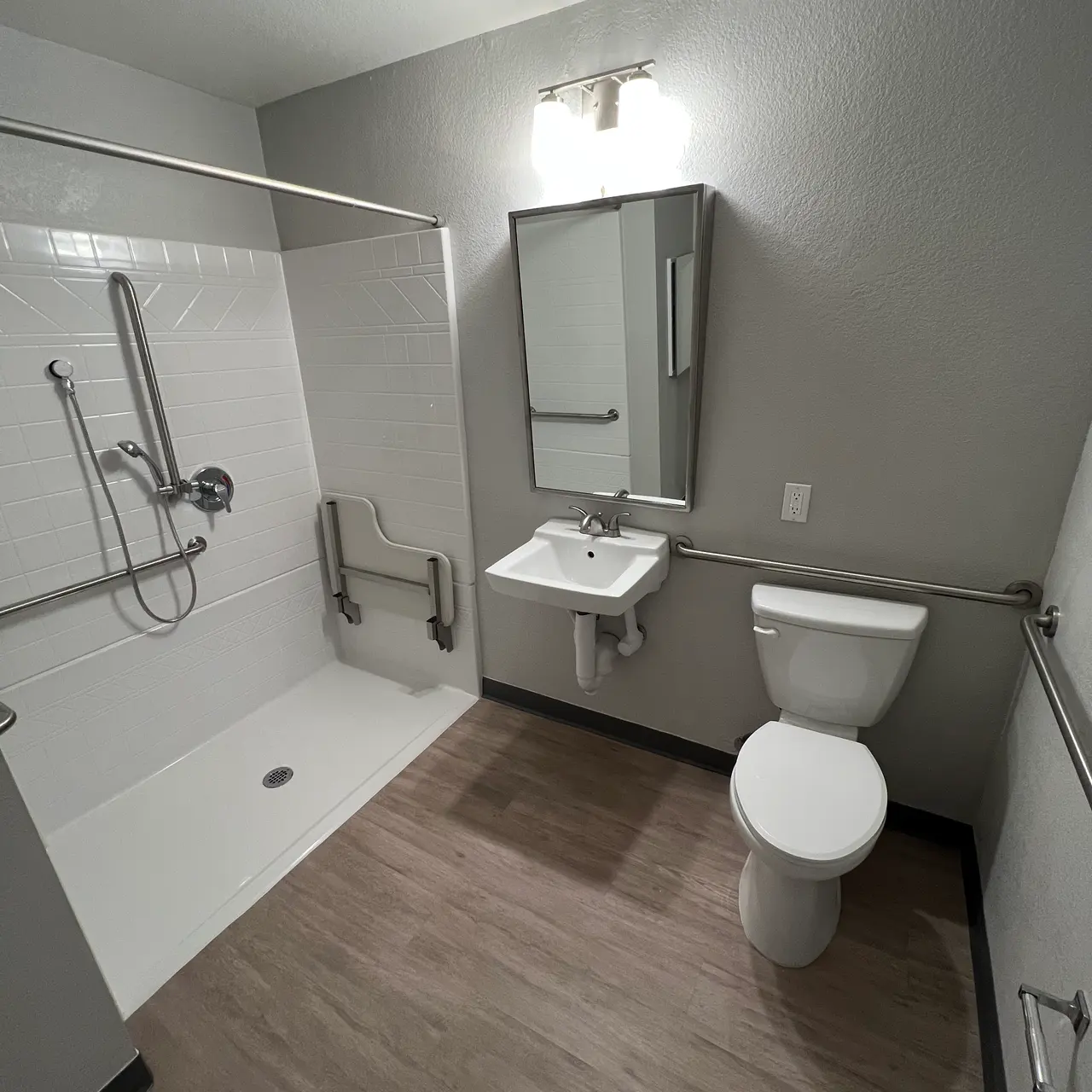 A clean, spacious bathroom featuring a walk-in shower with grab bars, a wall-mounted sink, and a toilet. The walls are painted in a light gray color and the floor is made of laminate wood. The lighting is soft and there is a mirror above the sink.