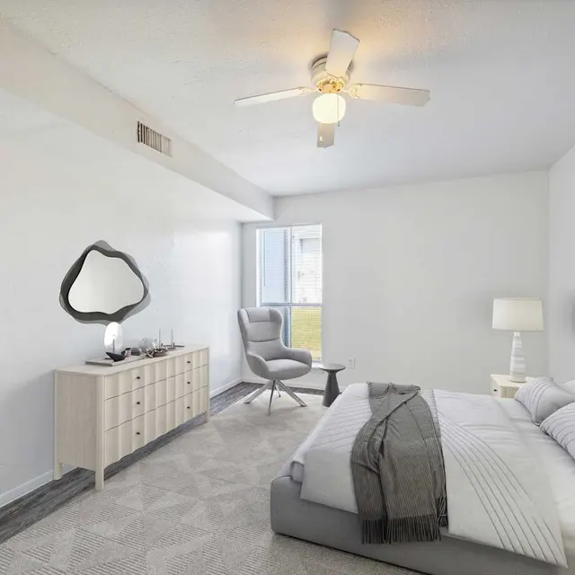 A modern bedroom featuring a neatly made bed with gray and white bedding, a side table with a lamp, a dresser with a decorative mirror, and a window allowing natural light to enter the space. The room has a minimalist aesthetic with light-colored walls and soft textures.