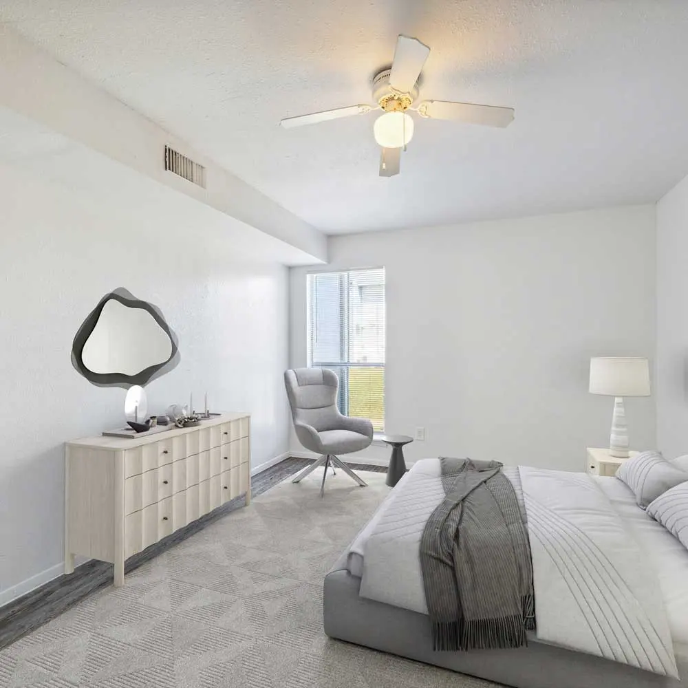 A modern bedroom featuring a neatly made bed with gray and white bedding, a side table with a lamp, a dresser with a decorative mirror, and a window allowing natural light to enter the space. The room has a minimalist aesthetic with light-colored walls and soft textures.