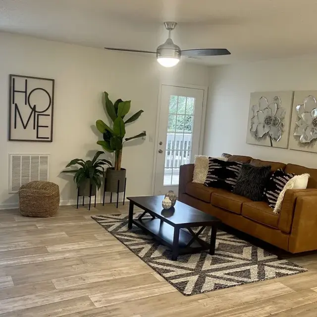 A modern living room featuring a brown leather sofa with decorative pillows, a black coffee table, and a geometric area rug. There is a tall plant in the corner, framed wall art with the word 'HOME', and large artworks on the wall. Natural light floods in through the windows, and a door leads to an outdoor area.