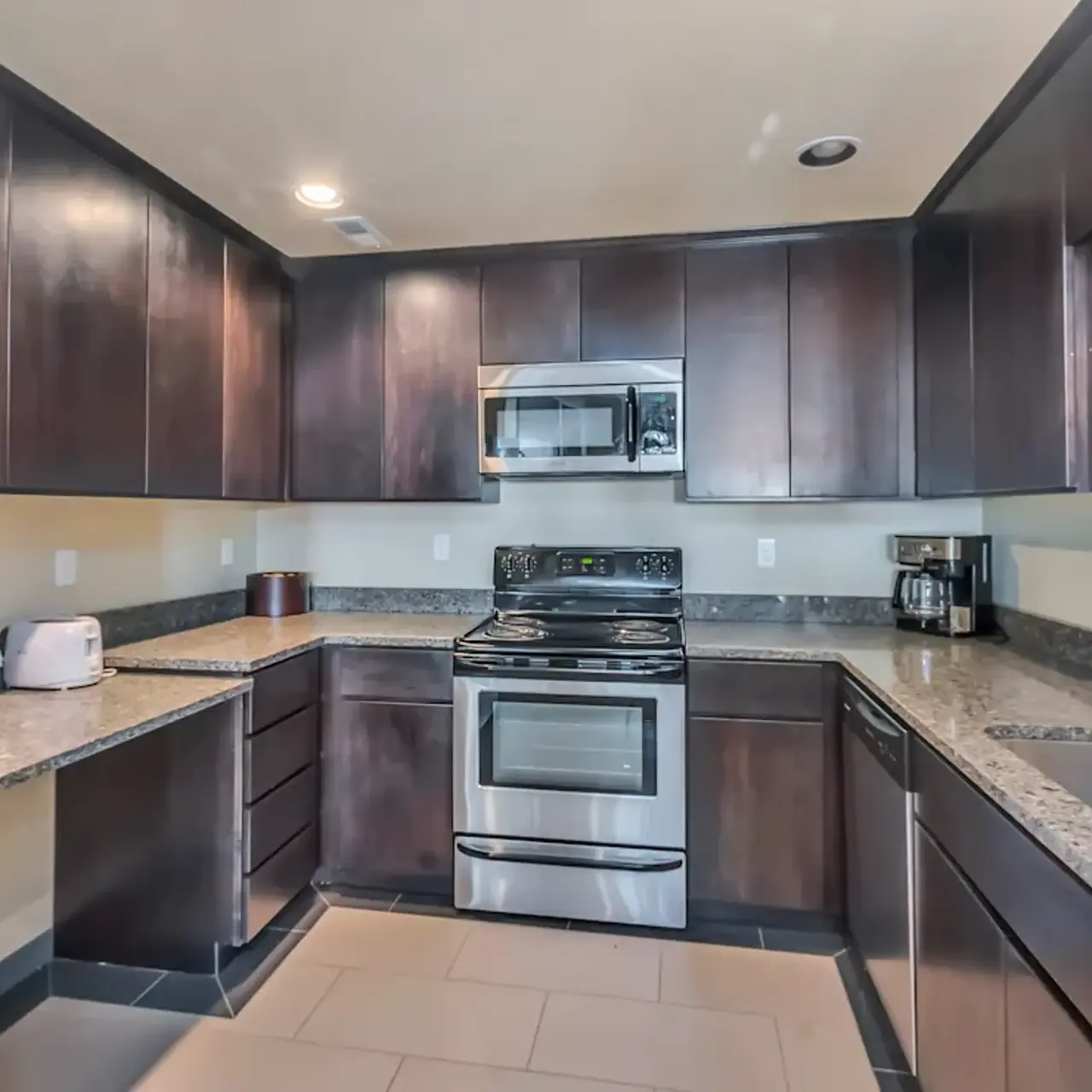 A modern kitchen featuring dark wood cabinets, a stainless steel refrigerator, a stainless steel stove, and granite countertops, with an open layout and ample storage.