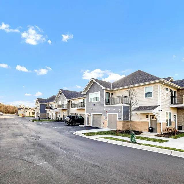 A modern apartment complex with two-story buildings situated along a recently paved road, surrounded by open space and blue sky.