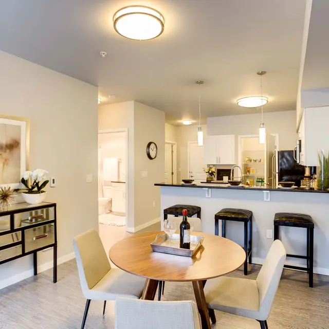 A stylish modern dining area and kitchen featuring a round table, four chairs, and a bar with stools. A decorative cabinet is placed against the wall, and there are plants and a framed artwork on display.