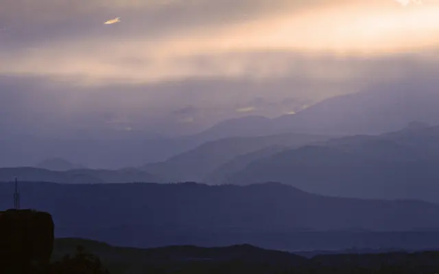 A panoramic view of layered mountains against a twilight sky, with soft hues of blue and purple.