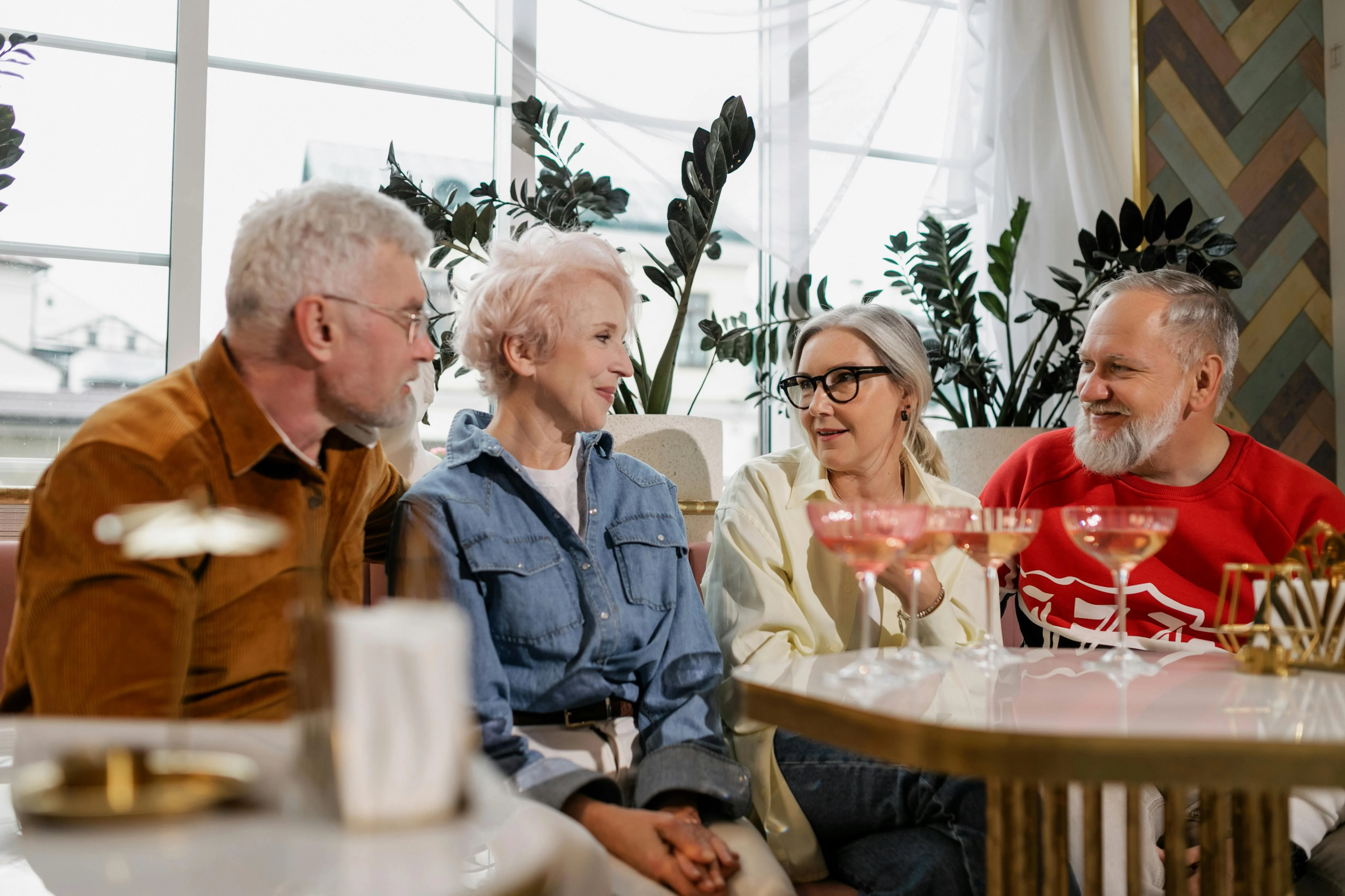 Elderly Friends Gathering A group of four elderly friends enjoying drinks at a stylish indoor setting with plants and cozy decor.