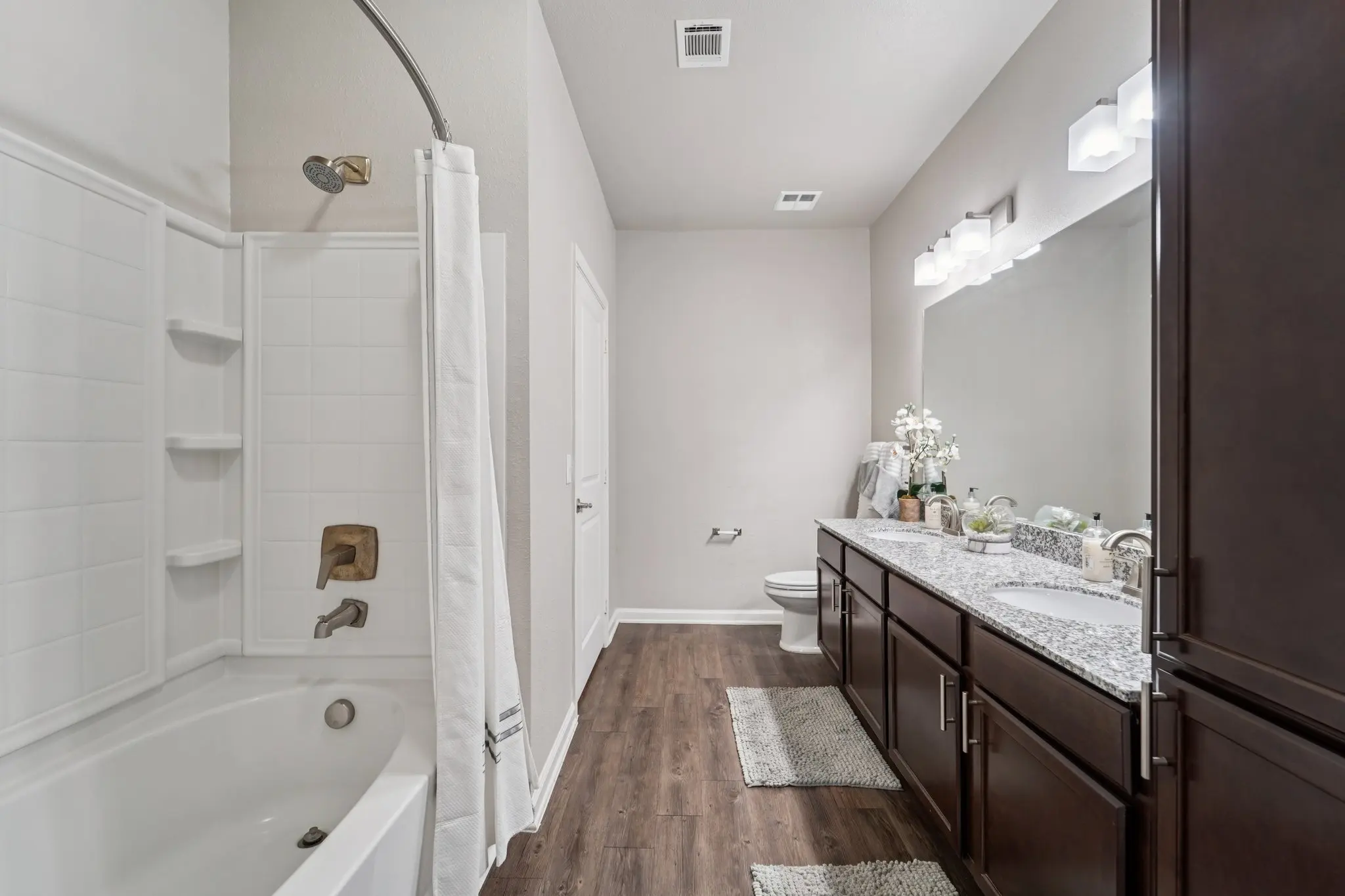 A spacious modern bathroom featuring a white bathtub with a shower, dark wood cabinets, a large mirror, and a granite countertop with toiletries. The flooring is dark wood, and the walls are painted light neutral shades.