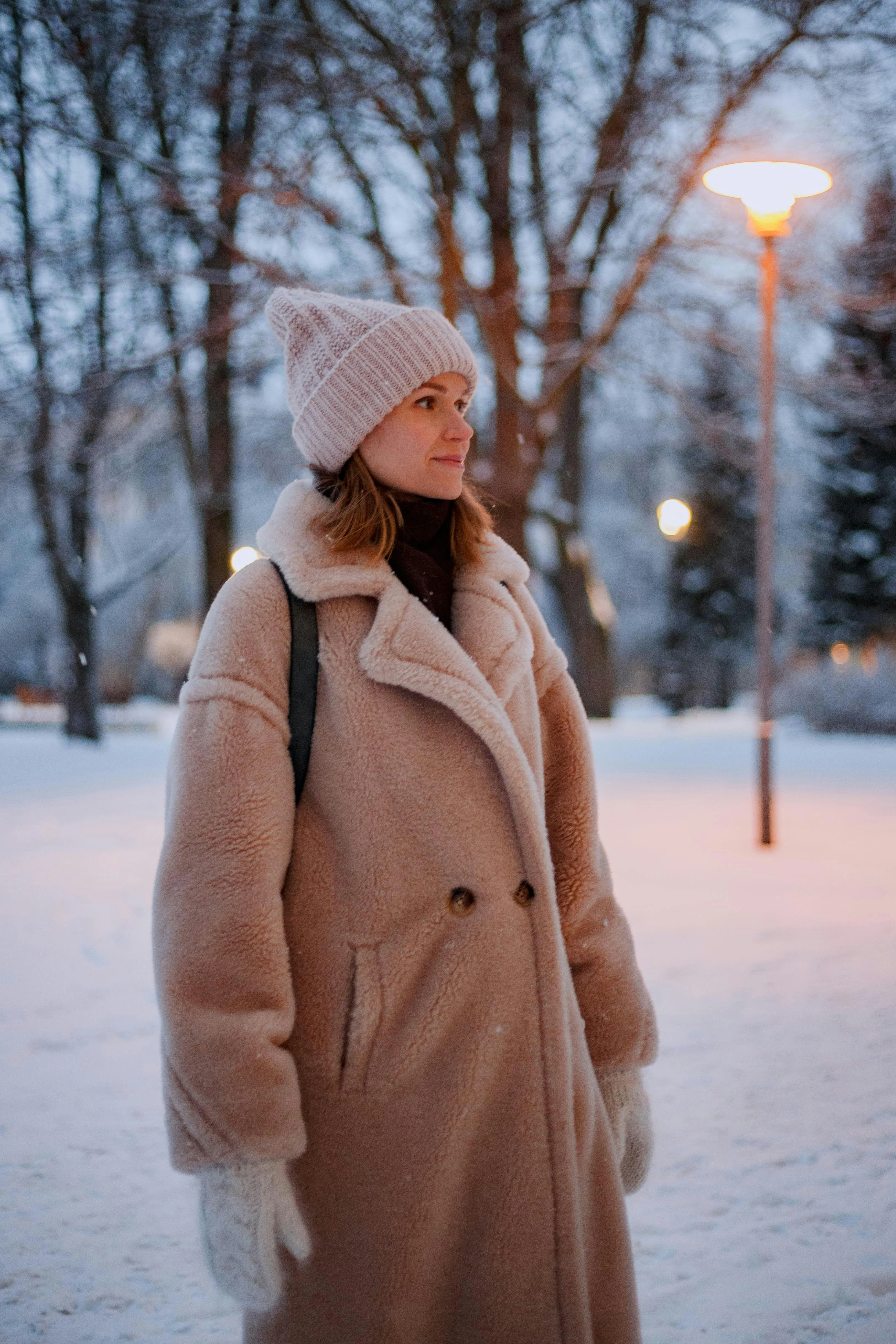 A woman wearing a cozy winter coat and hat stands outdoors in a snowy park during twilight. The surrounding trees are bare and a street lamp is illuminated in the background.
