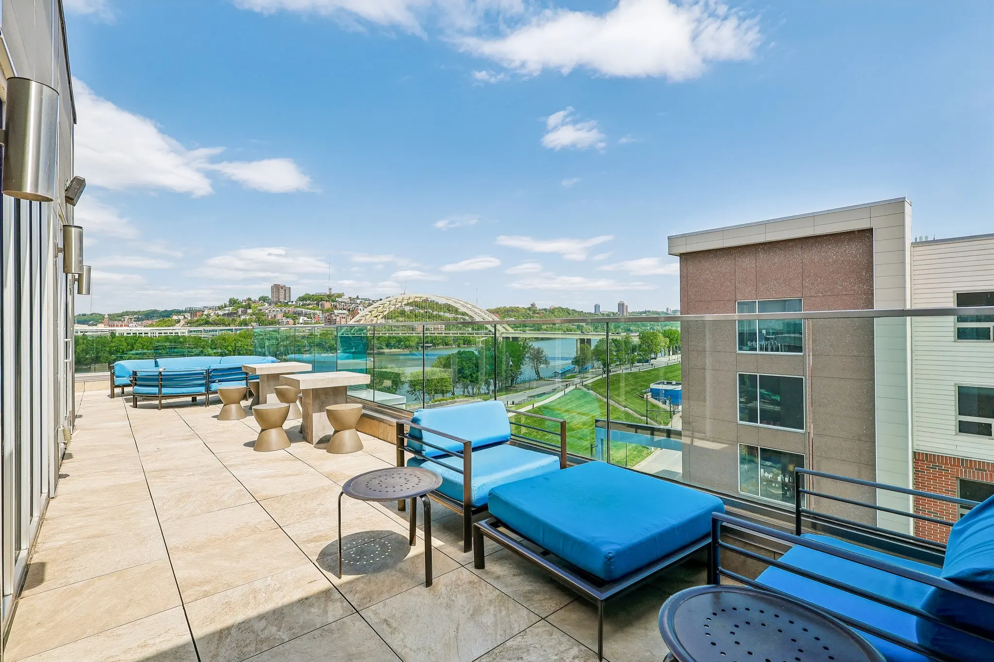 Rooftop Terrace with City Views A spacious rooftop terrace with blue lounge chairs and tables overlooking a green landscape and city skyline under a bright blue sky.