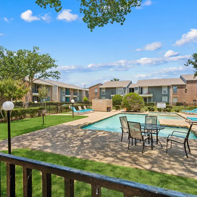 A view of a sunny apartment complex pool area with green grass, lounge chairs, and flowering bushes.