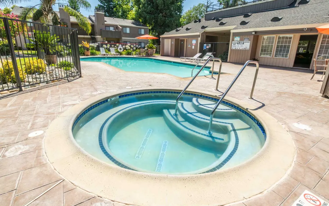 A view of a pool area featuring a jacuzzi with steps leading into the water, surrounded by lounge chairs and lush greenery.