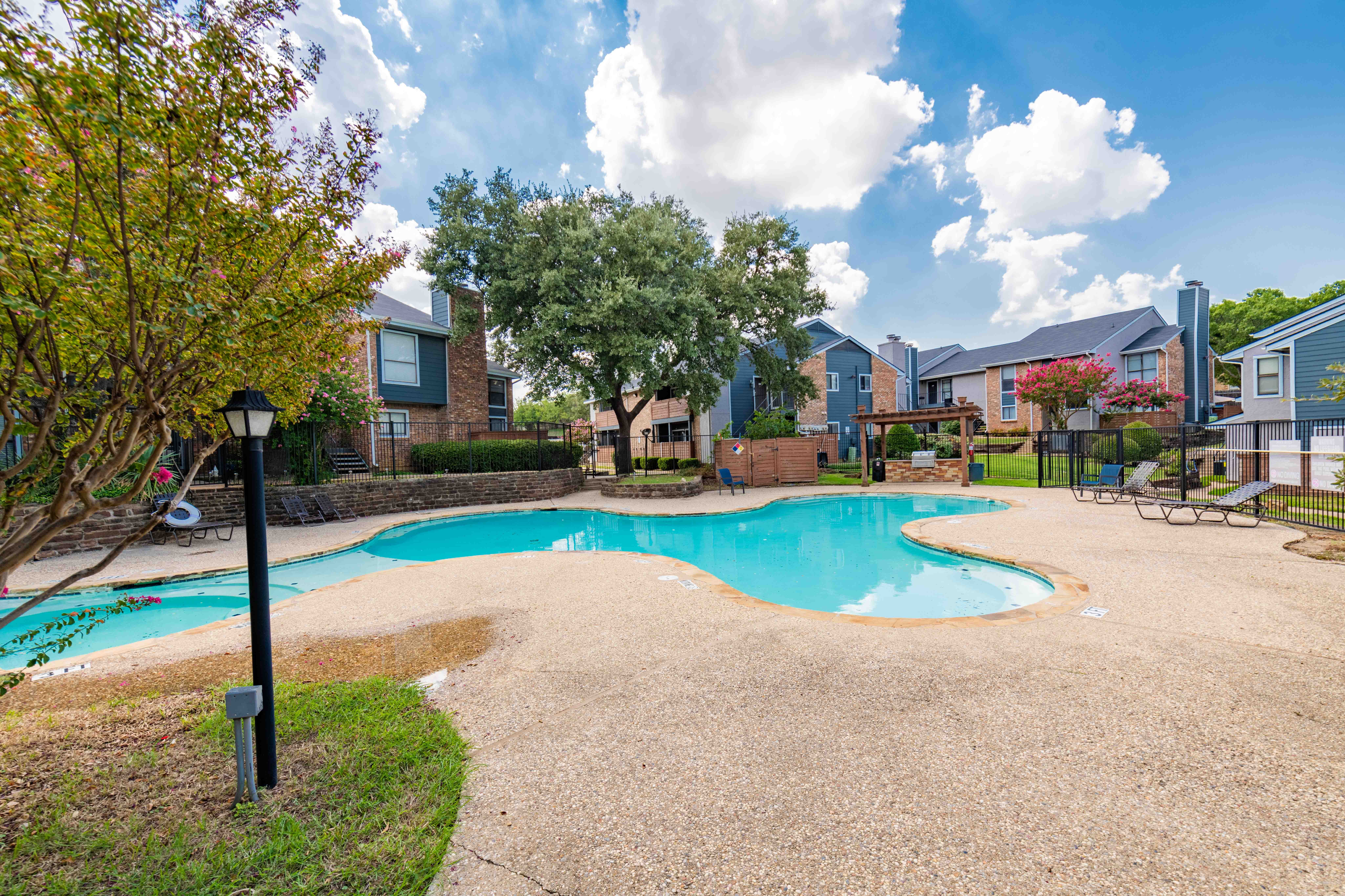 A view of a swimming pool surrounded by landscaped grounds, with nearby apartment buildings in the background and trees providing shade.