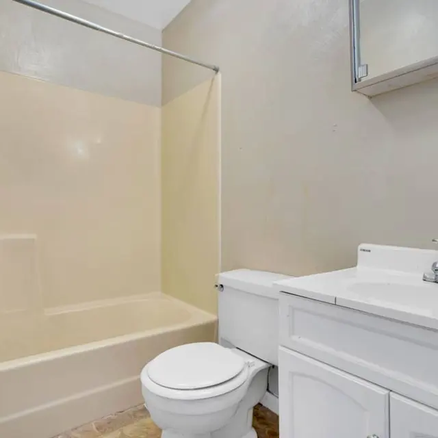 A clean and simple bathroom featuring a bathtub shower combination, a white toilet, and a white vanity with a sink. The walls are painted in a light color, and there is a mirror mounted above the vanity.