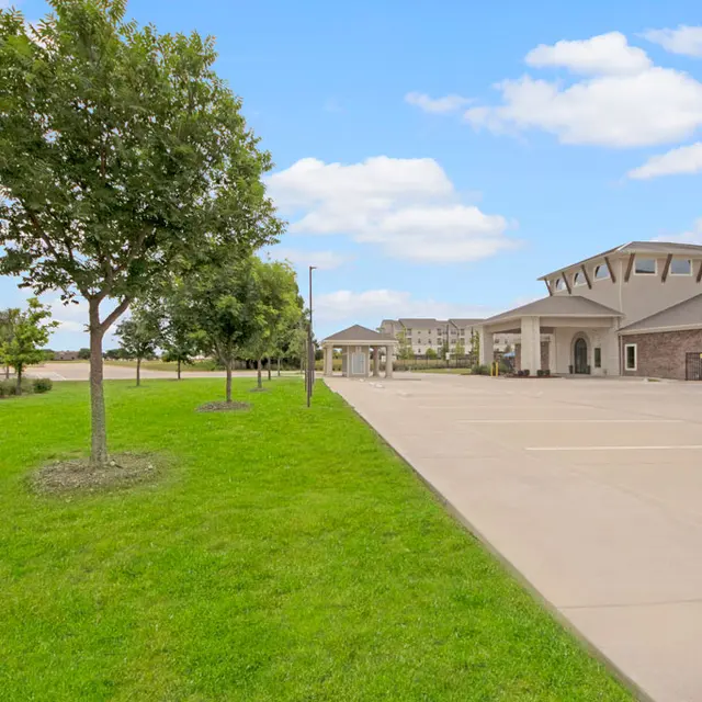 Community Entrance View A landscaped area with green grass and trees leading up to a modern building with a covered entrance. The sky is clear with a few clouds.