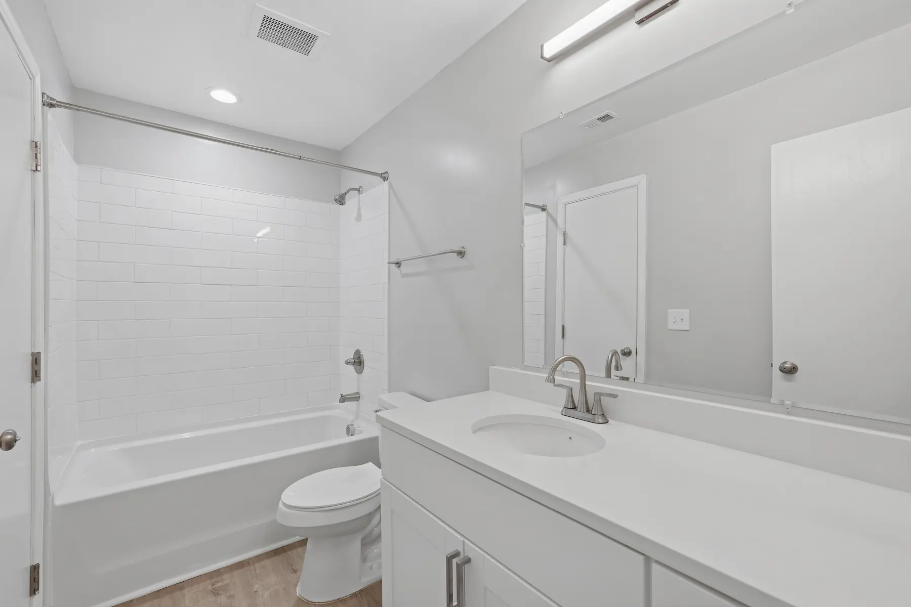 A clean, modern bathroom featuring a white bathtub, shower, toilet, and double vanity with a large mirror. The walls are painted in a light gray tone, enhancing the spacious feel of the room.