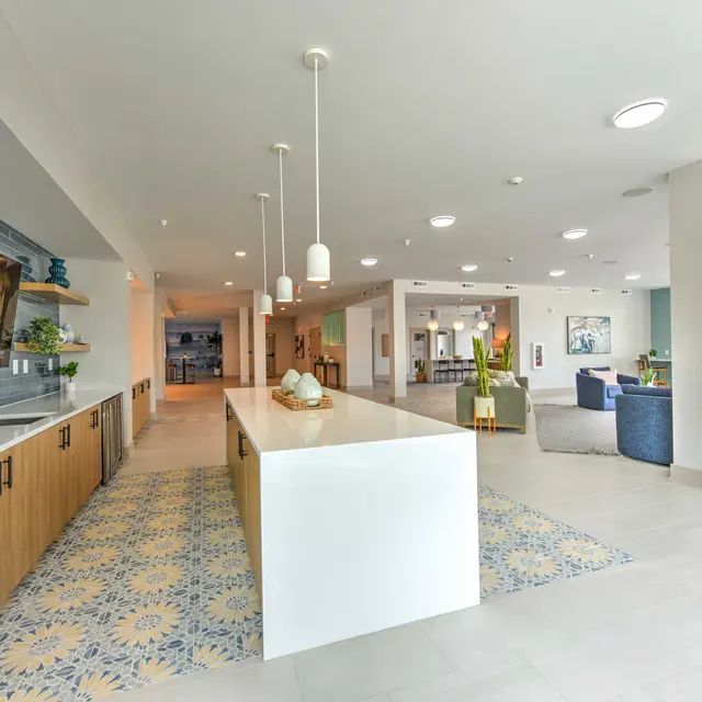 A spacious modern interior featuring a white countertop island, wooden cabinets, and a blue tiled wall with a television. The area has abundant natural light and a patterned floor rug.