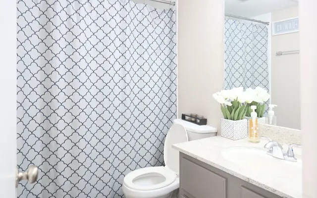 A modern bathroom featuring a white toilet, gray cabinetry, and a patterned shower curtain with navy blue designs. The counter has decorative items including flowers and toiletries.