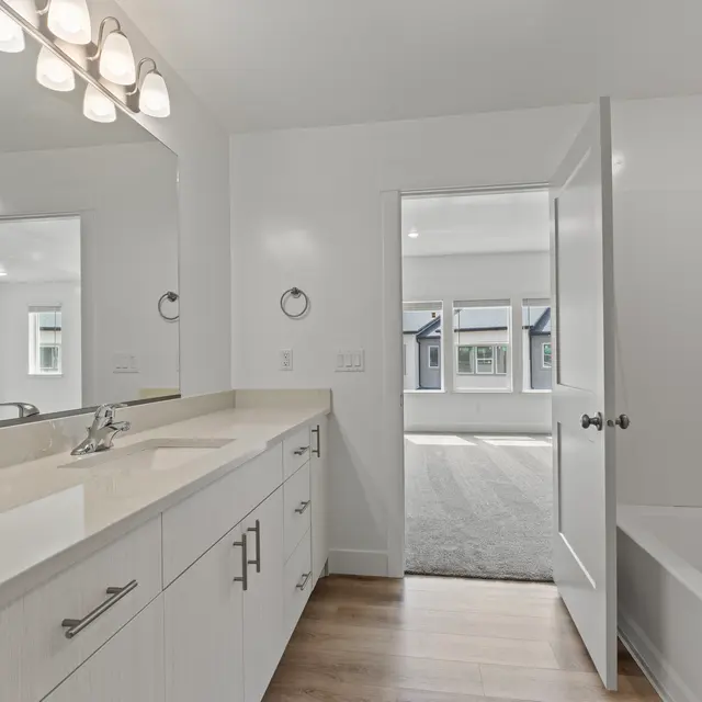A modern bathroom featuring a long countertop with a sink, white cabinetry, and a doorway leading to a spacious room with windows.