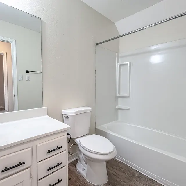 A modern bathroom featuring a white bathtub with a shower, a toilet, and a vanity with a mirror and drawers. The walls are light-colored, and there is a door leading to another room.